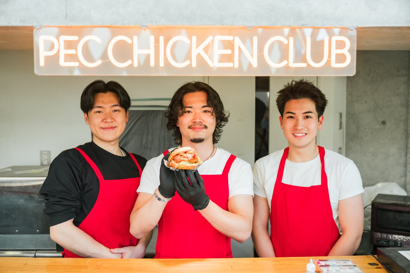 ネオンサイン「PEC CHICKEN CLUB」の前に、赤いエプロン姿の3人が並ぶ。真ん中の人がチキンバーガーを掲げている。